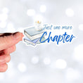 Load image into Gallery viewer, Love to read? If so then you know that there's always time for one more chapter! This individual die-cut sticker features a stack of books with a candle on top, and the words "One More Chapter", all in blue and gray on white. Enjoy your reading! This image shows a hand holding the Just One More Chapter sticker.