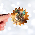 Load image into Gallery viewer, This image shows a hand holding the steampunk dragon on a gear sticker.