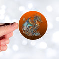 Load image into Gallery viewer, This image shows a hand holding the steampunk dragon copper background sticker.