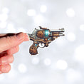Load image into Gallery viewer, This image shows a hand holding the Pirate Steampunk Pistol 2 Die-Cut Sticker.