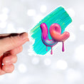 Load image into Gallery viewer, This individual die-cut sticker is one you either like or don't like; it's very unique! Featuring a 3D paint drip in pinks and purples over a green and blue paint swath, this sticker is hard to explain but pretty interesting to see. This image shows a hand holding the 3D Drip die-cut sticker.