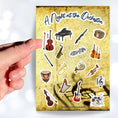 Load image into Gallery viewer, This sticker sheet celebrates all things music! Strings, brass, woodwinds, and percussion stickers that are perfect for any music lover. This image shows a hand holding a violin above the sticker sheet.