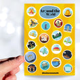Load image into Gallery viewer, Our Around the World sticker sheet collection has sticker images of iconic travel destinations! This sheet has a yellow background with 20 different stickers. This image shows a hand holding a sticker of ancient ruins with a night starry background above the sticker sheet.