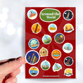 Load image into Gallery viewer, Our Around the World sticker sheet collection has sticker images of iconic travel destinations! This sheet has a red background with 18 different stickers. This image shows a hand holding a sticker of a night skyline above the sticker sheet.