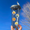 Load image into Gallery viewer, Our Around the World sticker sheet collection has sticker images of iconic travel destinations! This sheet has a red background with 18 different stickers. This image shows a water bottle with stickers of the Golden Gate Bridge, a lighthouse, a suspension bridge, and the Sidney Opera House applied to it.