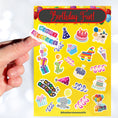 Load image into Gallery viewer, This birthday sticker sheet has a sparkle overlay with stickers showing all your favorite birthday celebration items like: Birthday cakes, presents, balloons, a piñata, birthday hats, and streamers. This image is of a hand holding one of the stickers above the sticker sheet.