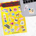 Load image into Gallery viewer, This birthday sticker sheet has a sparkle overlay with stickers showing all your favorite birthday celebration items like: Birthday cakes, presents, balloons, a piñata, birthday hats, and streamers. This image is of the sticker sheet next to an open laptop with 2 of the stickers applied to it.