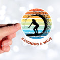 Load image into Gallery viewer, Hang 10 Bro! Catch a wave with this individual die-cut sticker featuring the silhouette of a female surfer inside the tube, all on a colored gradient background. Cowabunga! This image shows a hand holding the Catching a Wave die-cut sticker.