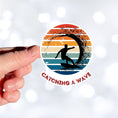 Load image into Gallery viewer, Hang 10 Bro! Catch a wave with this individual die-cut sticker featuring the silhouette of a male surfer inside the tube, all on a colored gradient background. Cowabunga! This image shows a hand holding the Catching a Wave sticker.