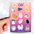 Load image into Gallery viewer, This sticker sheet is purrfect for all cat lovers! It has stickers of 12 different cute and cuddly cats, plus two pawprint stickers. This image shows a hand holding a brown cat sticker above the full sticker sheet.