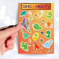 Load image into Gallery viewer, These dinosaur stickers are a-roar-able! This sticker sheet has all of your favorite dinosaurs plus dino eggs, plants, and dino footprints. This image shows a hand holding a tetradactyl sticker over the sticker sheet.
