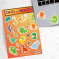 Load image into Gallery viewer, These dinosaur stickers are a-roar-able! This sticker sheet has all of your favorite dinosaurs plus dino eggs, plants, and dino footprints. This image has the sticker sheet next to an open laptop with a sticker of a dino hatching from and egg and a T-Rex dino sticker applied below the keyboard.