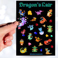 Load image into Gallery viewer, This sticker sheet is filled with cute images of baby dragons, and of course their treasure! This image shows a hand holding a sticker of a purple dragon sitting with butterflies flying around over the sticker sheet.