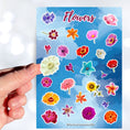Load image into Gallery viewer, So many flowers! This sticker sheet is filled with images of all your favorite flowers. This image shows a hand holding a white flower above the sticker sheet.