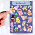Load image into Gallery viewer, Who doesn't love gnomes? This sparkle sticker sheet features a host of gnomes along with rainbows, stars, balloons, and other sticker images. All with a sparkle holographic overlay! This image shows a hand holding a yellow gnome over the sticker sheet.