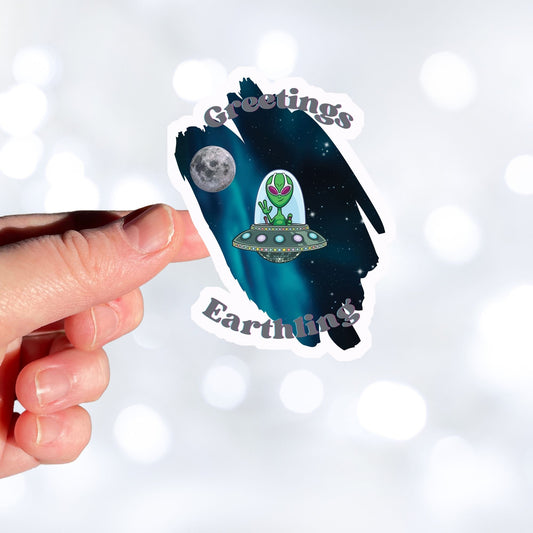 Welcome space aliens! This individual die-cut sticker features a friendly green space alien in a flying saucer, on a background of stars with the moon behind, and the words "Greetings Earthling". This image is of a hand holding the flying saucer sticker.