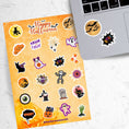 Load image into Gallery viewer, Ghosts, skeletons, black cats, and spiders; this spooky but fun sticker sheet is perfect for your Halloween decorating! This image shows the sticker sheet next to an open laptop with a sticker of bats flying in front of the moon, and a sticker that says "Boo!" on a black background applied below the keyboard.