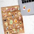 Load image into Gallery viewer, Cheers! Filled with sticker images of your favorite cocktails and adult beverages, this is for adults who can enjoy this sticker sheet responsibly! This image shows the sticker sheet next to an open laptop with stickers of mixed drinks applied below the keyboard.