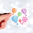 Load image into Gallery viewer, Float away on a colorful hot air balloon! This individual die-cut hot air balloon sticker has six, yes six, different hot air balloons on a background of clouds. This image shows a hand holding the hot air balloon sticker.
