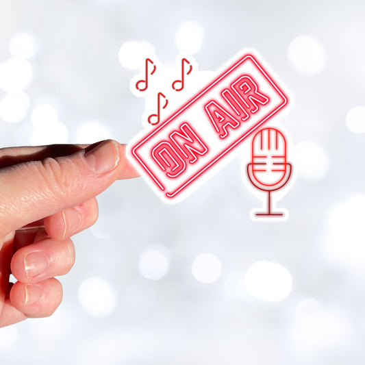 Music, TV, Podcast, or even Amateur Radio - this sticker is great for anyone who is On Air! This individual die-cut sticker is in neon red with the words On Air above a microphone with music notes above. Please note that the large version of this sticker is on a white background and cut around the image, and the small version is on a black circle. This image shows a hand holding the large On Air sticker.