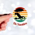 Load image into Gallery viewer, Who doesn't want to be on vacation? Whether you travel or just stay around home, being on vacation sure beats the alternative! This individual die-cut sticker features the silhouette of a beach chair and umbrella with a cool tropical beverage easily at hand, all on a colored gradient background. Relax, you're On Vacation! This image shows a hand holding the On Vacation sticker.