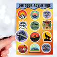 Load image into Gallery viewer, Tie up your boots and enter the wild with this sticker sheet! This sticker sheet has twelve different stickers showing different outdoor activities. From the mountains to camping to climbing this set is perfect for any outdoors person. This image shows a hand holding a sticker showing a climber on a mountain that says "Climbing".
