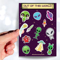 Load image into Gallery viewer, This sticker sheet is out of this world with individual stickers of aliens, space ships, planets, and alien creatures. This image is of a hand holding one of the individual stickers above the sticker sheet