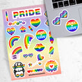 Load image into Gallery viewer, Show your LGBTQ Pride with this sticker sheet! This sticker sheet is filled with rainbow Pride images that are perfect for anywhere you want to show your Pride. This image is of the sticker sheet next to an open laptop with a sticker of heart rainbow glasses and an sticker saying Just Love applied below the keyboard.