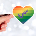 Load image into Gallery viewer, This individual die-cut sticker features a heart shape with graduated colors from orange at the top to blue at the bottom, and the word love written across. Perfect for the people you love! This image shows a hand holding the Love Heart sticker.