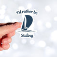 Load image into Gallery viewer, This individual die-cut sticker is great for anyone who loves to sail! It features a blue sailboat with the words "I'd rather be Sailing". This sailing sticker is understated, but says a lot! This image shows a hand holding the I'd rather be Sailing sticker.