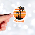 Load image into Gallery viewer, Grab your favorite beverage and relax under the palm trees! This individual die-cut sticker features two palm trees and an umbrella drink in a coconut shell, on an orange and white gradient background, with the word "Relaxing" below. This image shows a hand holding the relaxing sticker.