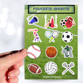 Load image into Gallery viewer, What are your favorite sports? Football, gymnastics, soccer, volleyball, cheer, baseball, hockey, tennis, or lacrosse? If you love any of these then this sticker sheet is perfect for you! This image shows a hand holding a baseball sticker above the Favorite Sports sticker sheet.