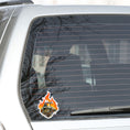 Load image into Gallery viewer, This individual die-cut steampunk sticker features a brown engine with a cutaway to show the gears inside, on a background of yellow, orange, and red flames. This image shows the steampunk engine sticker on the back window of a car.