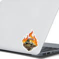 Load image into Gallery viewer, This individual die-cut steampunk sticker features a brown engine with a cutaway to show the gears inside, on a background of yellow, orange, and red flames. This image shows the steampunk engine sticker on the back of an open laptop.
