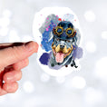 Load image into Gallery viewer, Steampunk Rotty is a happy looking Rottweiler with a steampunk hat and goggles, all on a pastel background. This image shows a hand holding the steampunk rotty sticker.