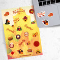 Load image into Gallery viewer, This festive Thanksgiving sticker sheet is perfect for decorating or scrapbooking. It features all of your favorite Thanksgiving images and inspirational sayings. Let the feast begin! This image shows the Happy Thanksgiving sticker sheet next to a laptop with stickers of pumpkin and pumpkin pie, a turkey dressed as a pilgrim, and the words "Let's Feast" applied below the keyboard.