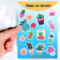 Load image into Gallery viewer, Let your green thumb shine with this sticker sheet. It features a variety of succulent plant stickers that are perfect for your favorite gardener! This image shows a hand holding a potted succulent in a green and blue container sticker held above the sticker sheet.
