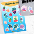 Load image into Gallery viewer, Let your green thumb shine with this sticker sheet. It features a variety of succulent plant stickers that are perfect for your favorite gardener! This image shows the sticker sheet next to an open laptop with a round terrarium sticker and a potted succulent plant sticker, both in pastel colors, applied below the keyboard..