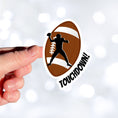 Load image into Gallery viewer, This individual die-cut sticker features the silhouette of a football (American) player about to pass the ball, on a football background, with the word "Touchdown!" on the lower right side. This image shows a hand holding the touchdown sticker.