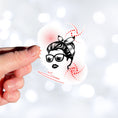 Load image into Gallery viewer, Trash Polka uses red, black, and white with a combination of abstract, surrealistic, and realistic images, and this individual die-cut sticker features a woman wearing sunglasses with her hair tied up on a white background with red swirls. This image shows a hand holding the trash polka woman sticker.