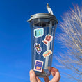 Load image into Gallery viewer, Pack your bags and grab your passport; it's travel time! This sticker sheet is filled with travel related sticker images sure to please even the most discerning traveler. Some of the sticker images are an airplane, suitcase, globe, and ship. This image is of a water bottle with stickers of a suitcase, a globe, a camera, a bus, and a travel stamp applied to it.
