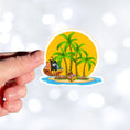 Load image into Gallery viewer, Pirate's Treasure awaits on this topical island! This individual die-cut sticker features an island with palm trees in front of the setting sun, and yes that is a treasure chest on the beach. But you'd better hurry before the pirate ship gets there and makes off with the prize! This image shows a hand holding the Treasure Island sticker.