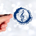 Load image into Gallery viewer, This individual die-cut sticker features a blue treble clef with a paint splattered background. Perfect for performers and music lovers alike! This image shows a hand holding the treble clef sticker.