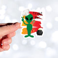 Load image into Gallery viewer, I wouldn't trust this space alien! This individual die-cut sticker features a green space alien flashing a peace sign, but wearing a chefs hat and trying to hide a wooden spoon, with a boiling pot behind, all on an orange background with the words "Trust Me!" in the upper right corner. This image shows a hand holding the trust me alien sticker.