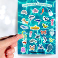 Load image into Gallery viewer, This sticker sheet is filled with fun and cute sticker images of all your favorite undersea creatures! Images include whales, sharks, jellyfish, manatee's, and crabs. There's even a yellow submarine! This image shows a hand holding a sticker of a blue and green seahorse above the sticker sheet.