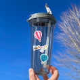 Load image into Gallery viewer, Fly away in a hot air balloon, a plane, or a helicopter with this sticker sheet. This image shows a water bottle with stickers of a red and white hot air balloon, a plan pulling a banner of multi-colored triangle flags, and a blue striped hot air balloon, applied to it.