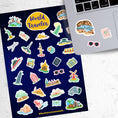 Load image into Gallery viewer, Our World Traveler sticker sheet has sticker images of iconic travel destinations! This sheet has a black background with over 30 different stickers including the Statue of Liberty, Easter Island, Stonehenge, and a passport and camera. This image shows the sticker sheet next to an open laptop with stickers of the Grand Canyon, sunglasses, and a travel notebook and pen, applied below the keyboard.
