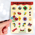 Load image into Gallery viewer, This sticker sheet has a sparkle overlay with 19 different stickers of cute ladybugs and bees. This image shows a hand holding a sticker of a ladybug with a flower over the sticker sheet.