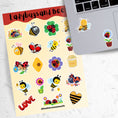 Load image into Gallery viewer, This sticker sheet has a sparkle overlay with 19 different stickers of cute ladybugs and bees. This image shows the sticker sheet next to an open laptop with a sticker of a bee mom with her baby bee and a sticker of a ladybug on a flower applied below the keyboard.