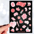 Load image into Gallery viewer, Let's celebrate mom! Mom's can do it all, and this pink aesthetic sticker sheet has sticker images of things that moms, and non-moms, enjoy like - candles, bubble baths, makeup, or coffee. This image shows a hand holding a sticker that says "Mom Life" in the shape of a heart above the sticker sheet.